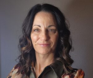 Close-up portrait of a smiling woman with dark wavy hair wearing a floral blouse against a gray wall.